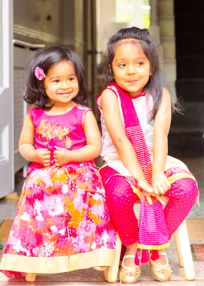 Two girls sitting on stools in Indian dresses in the doorway of their house in North London