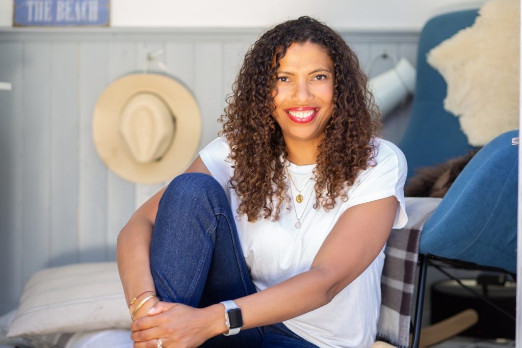Woman with long curly hair wearing jeans and sitting in beach hut. Blue chair behind her with dog sleeping